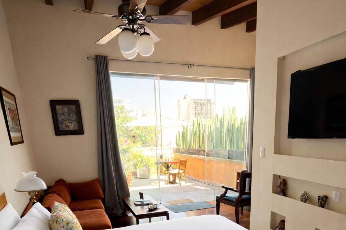 Habitación de hotel con una cama grande, ventilador de techo, balcón con mesa y sillas, y televisión montada en la pared.