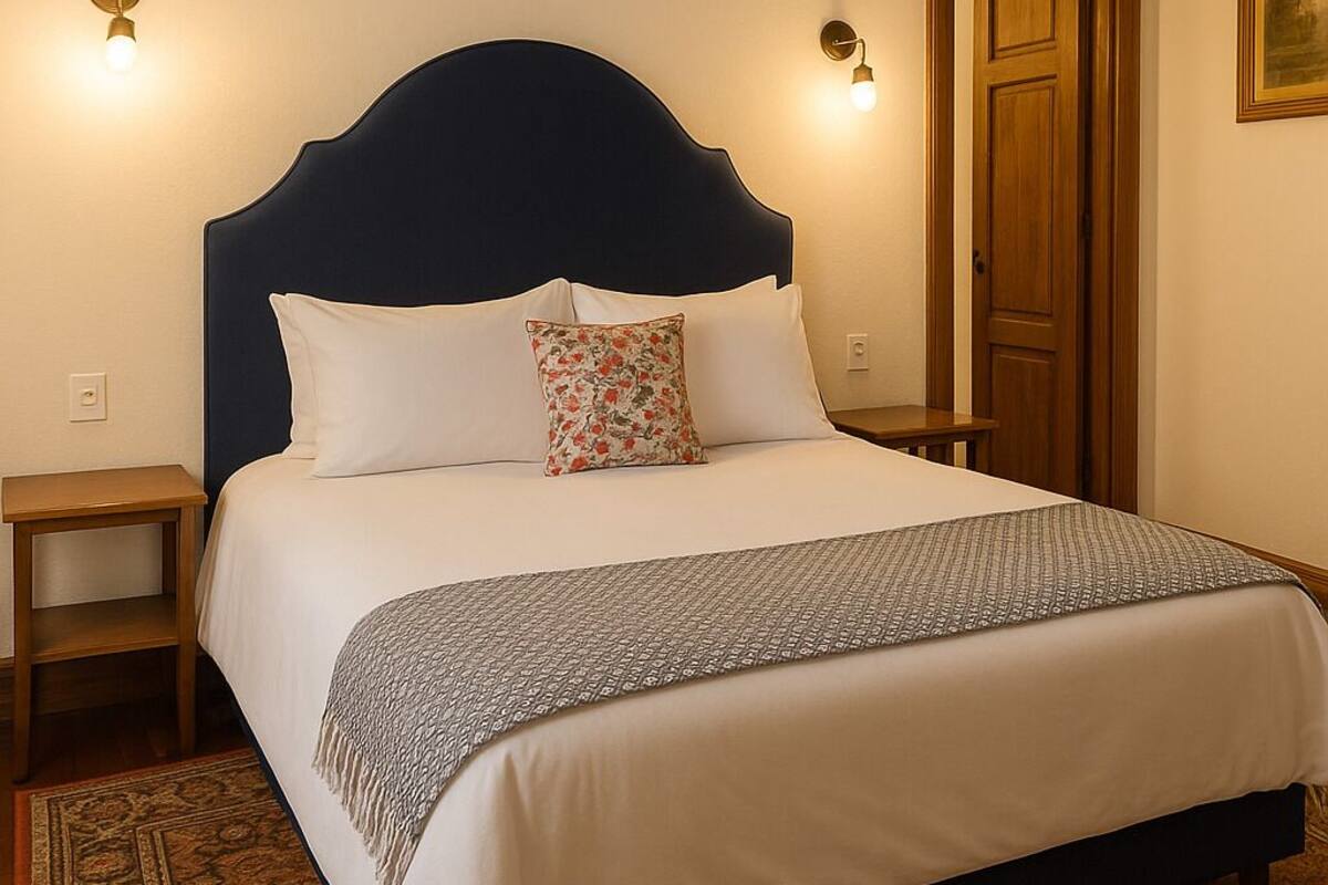 Una cama bien hecha con un cabecero oscuro, ropa de cama blanca y una manta estampada. Hay dos mesitas de noche con lámparas, una puerta de madera y una alfombra estampada en el suelo.