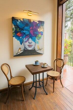 Une pièce dotée d’un plancher en bois, de deux fauteuils en osier, d’une petite table en bois sur laquelle se trouvent une tasse et une soucoupe, et d’un tableau abstrait aux couleurs vives accroché au mur.