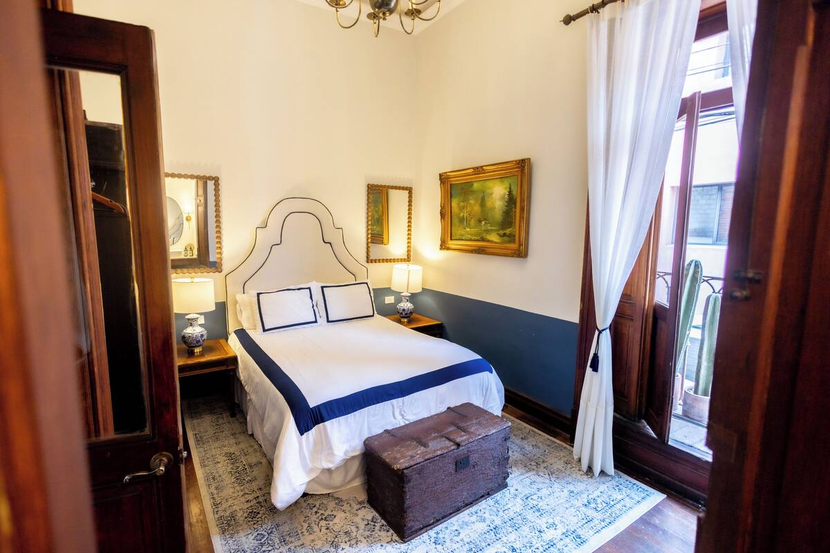 Un dormitorio con cama, lámparas de noche, una mesita de noche, un cofre de madera, una ventana con cortinas y un cuadro en la pared.