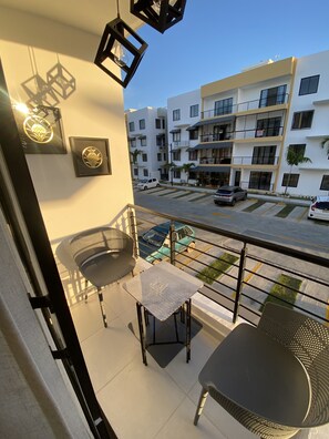 Un balcon avec deux chaises et une petite table, donnant sur une rue où sont garées des voitures et sur un immeuble à plusieurs étages en arrière-plan.