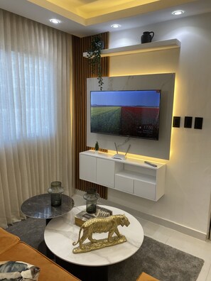 Un soggiorno moderno con una TV a schermo piatto montata su una parete bianca, un tavolino rotondo con una statuetta decorativa di un tigre e una piccola pianta in vaso sullo scaffale sopra la TV.