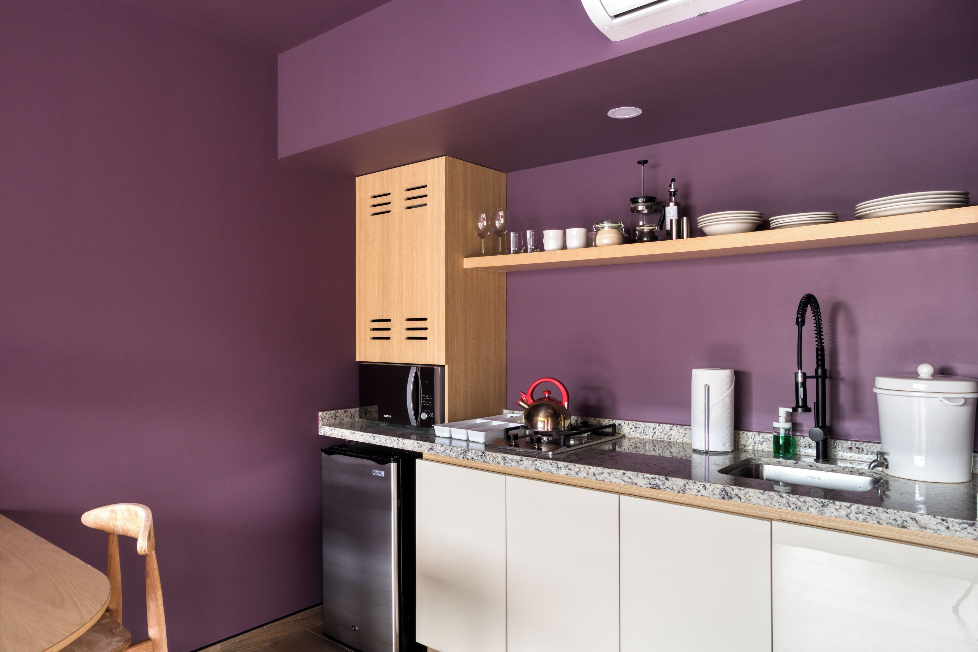 A modern kitchen with a purple wall, wooden cabinets, a stainless steel refrigerator, a sink, and a countertop with various kitchen items.