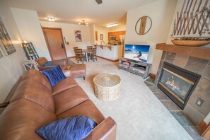 A living room with a brown leather sofa, a TV, a fireplace, and a kitchen in the background. - Oro Grande 308 (Keystone)