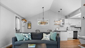 Ein modernes Wohnzimmer mit einer grauen Couch, gemusterten Kissen, einem schwarzen Couchtisch und einem Teppich mit Muster. Im Hintergrund ist ein Essbereich mit Stühlen und einer Chandelier zu sehen.