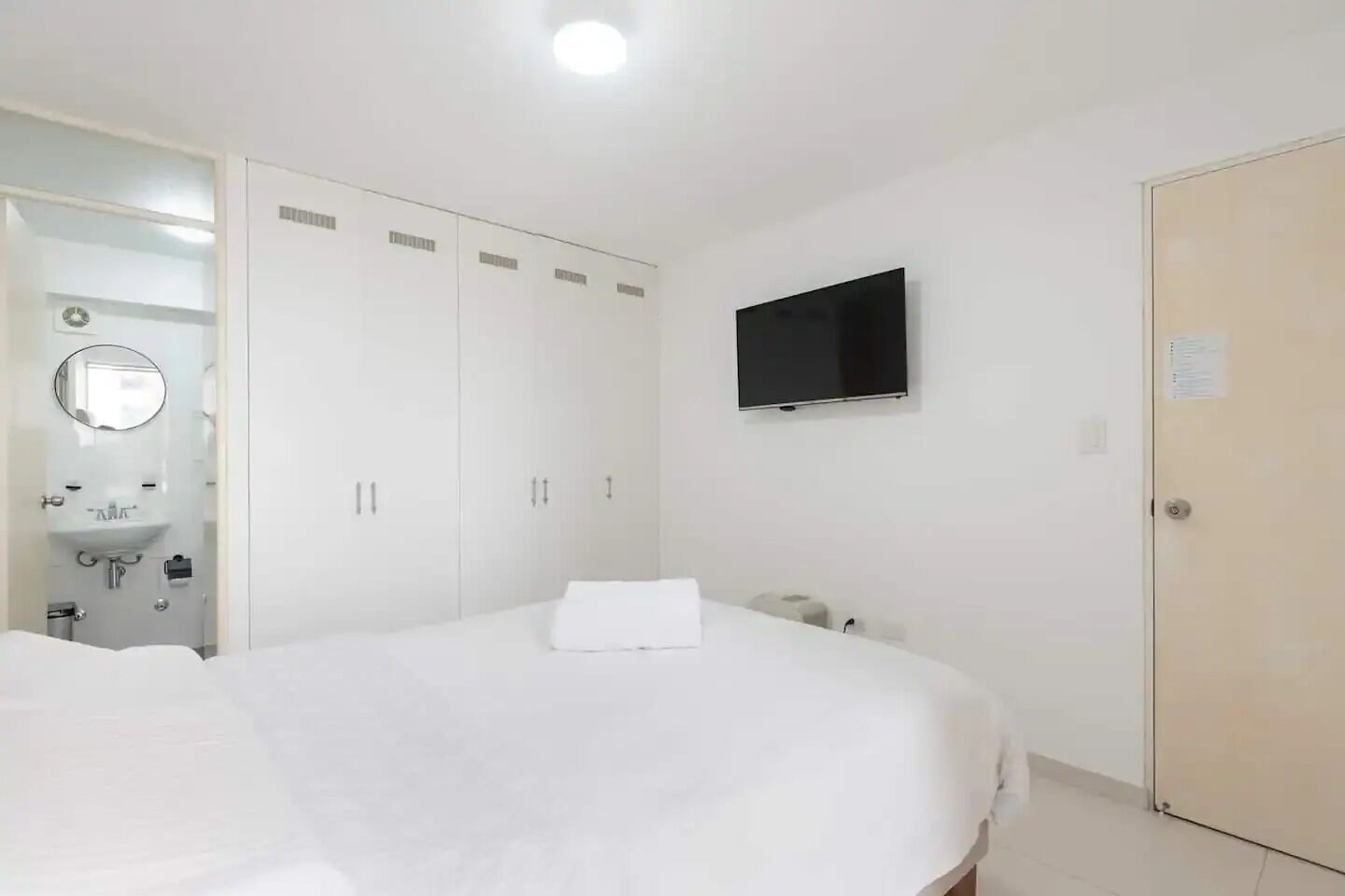 A hotel room with a bed, a TV mounted on the wall, a wardrobe, and a bathroom visible through an open door.