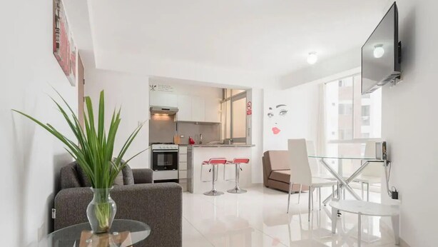 Un salon moderne avec un canapé, une table basse en verre et une plante en pot. On y trouve également un coin cuisine équipé d’une cuisinière, d’un réfrigérateur et d’une table à manger avec des chaises. Un téléviseur à écran plat est fixé au mur.