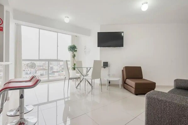 A modern living room with a sofa, a dining table with chairs, a TV mounted on the wall, and a balcony with a view of the city.