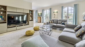 A modern living room with a flat-screen TV, grey sofas, a glass coffee table, and a large window with curtains. - Historic Villa 9 people, Private Pool, near Cannes (Grasse)
