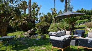 An outdoor seating area with wicker furniture and a large umbrella, surrounded by lush greenery and palm trees. - Historic Villa 9 people, Private Pool, near Cannes (Grasse)