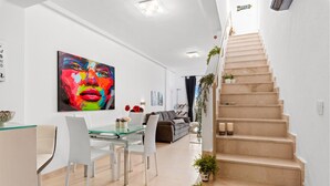 Un salon moderne avec une table à manger en verre, des chaises blanches, un canapé gris et un tableau abstrait aux couleurs vives. Un escalier orné de plantes en pot mène à un niveau supérieur.