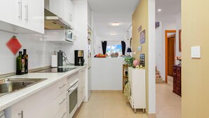 Une cuisine moderne avec des armoires blanches, un évier en acier inoxydable et un four à micro-ondes. On y trouve une fenêtre avec des rideaux, un réfrigérateur et une armoire en bois ornée d’une plante.