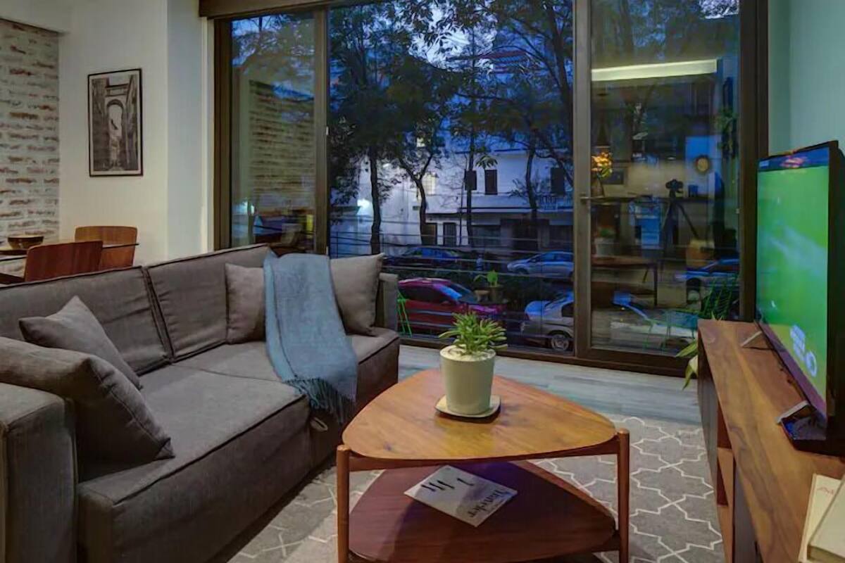 Un salon moderne avec un canapé, une table basse agrémentée d’une plante, un téléviseur et une grande fenêtre donnant sur la ville.
