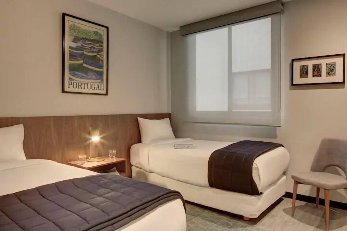 Une chambre d’hôtel avec deux lits, une fenêtre avec des stores, un tableau représentant un paysage du Portugal et une chaise.
