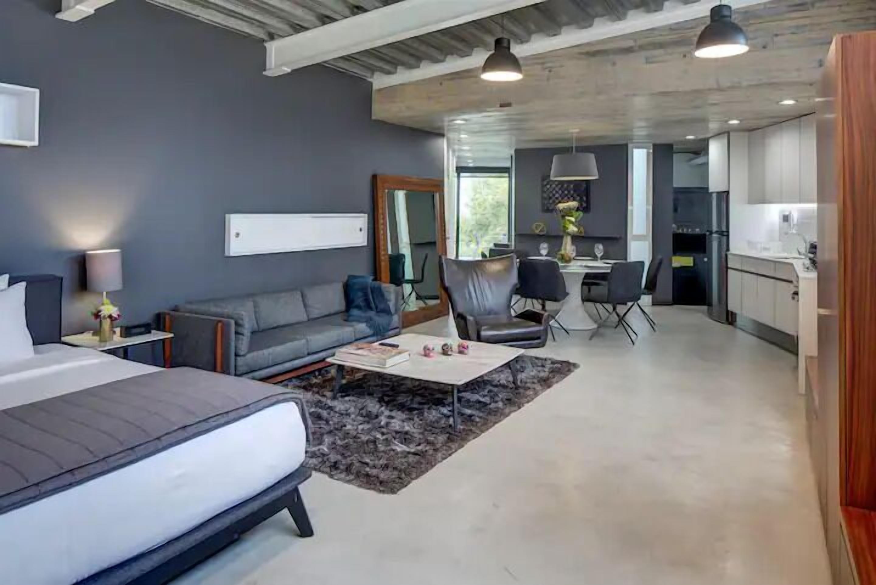A modern hotel room with a large bed, a gray sofa, a dining area with a table and chairs, and a kitchen in the background.