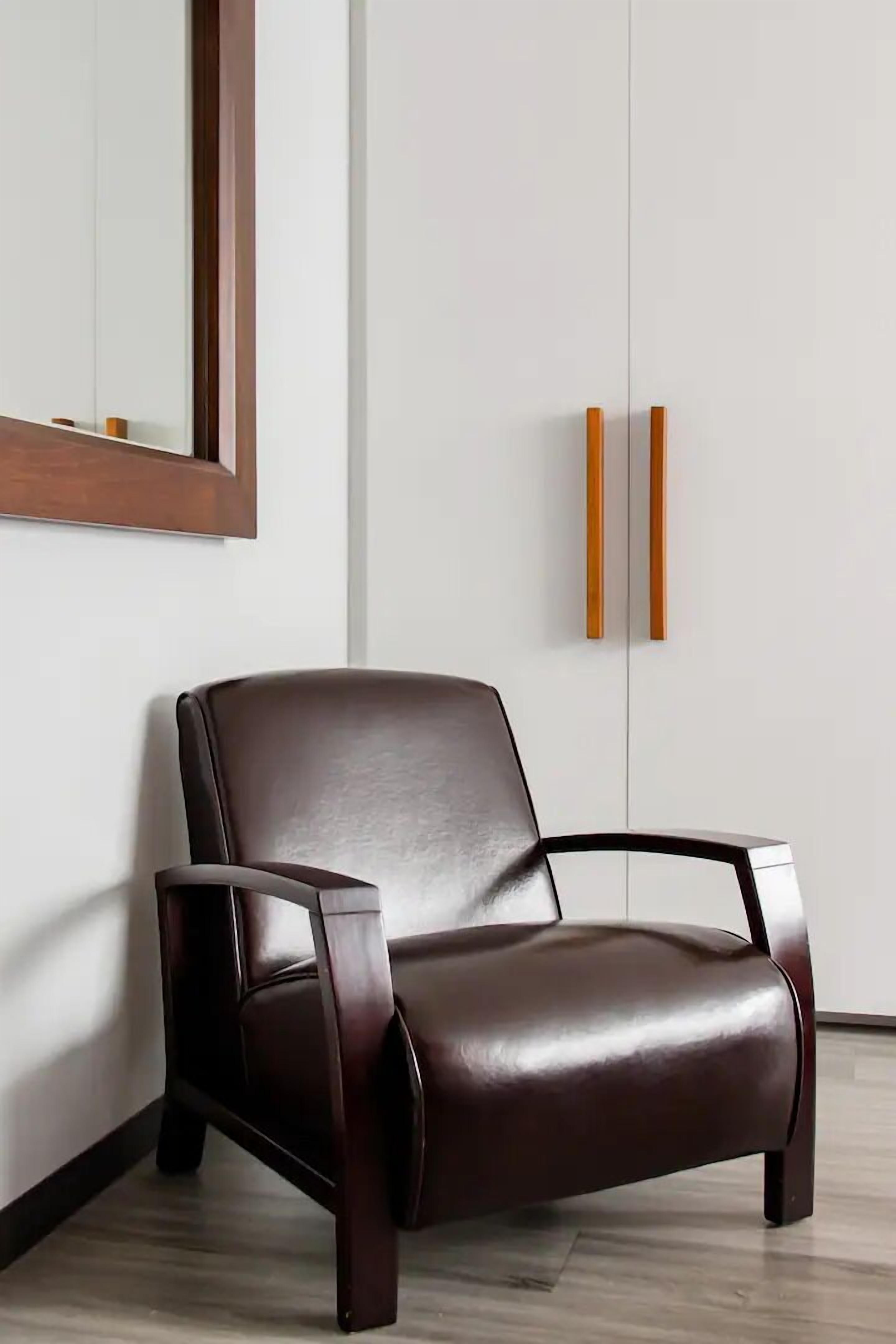 Un sillón de cuero marrón oscuro con patas y marco de madera, colocado frente a una pared blanca con un espejo de marco de madera arriba.