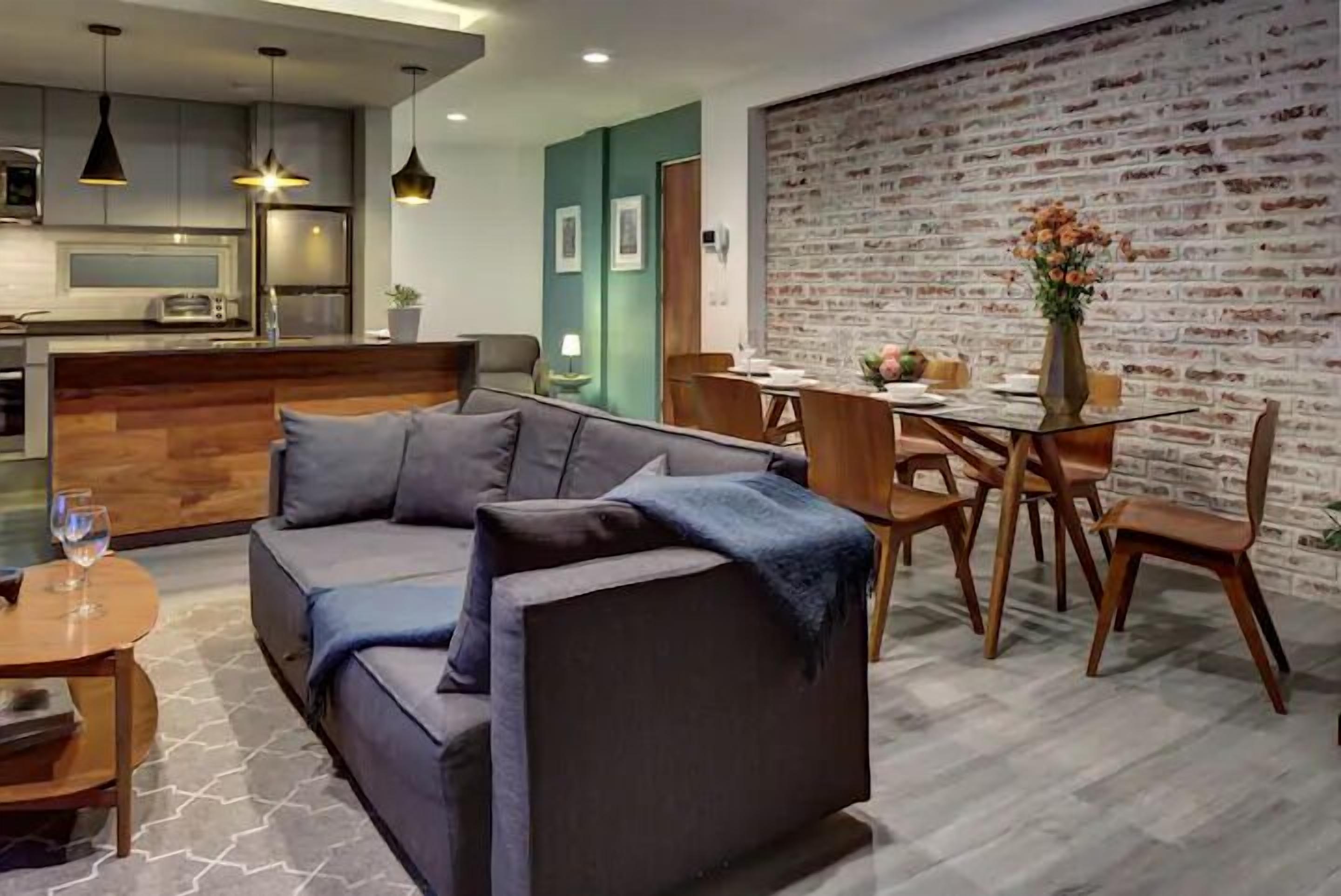 Een moderne zithoek met een bank, eettafel en stoelen. Er is een keuken met een koelkast en een magnetron. De kamer heeft een bakstenen muur en een houten vloer.