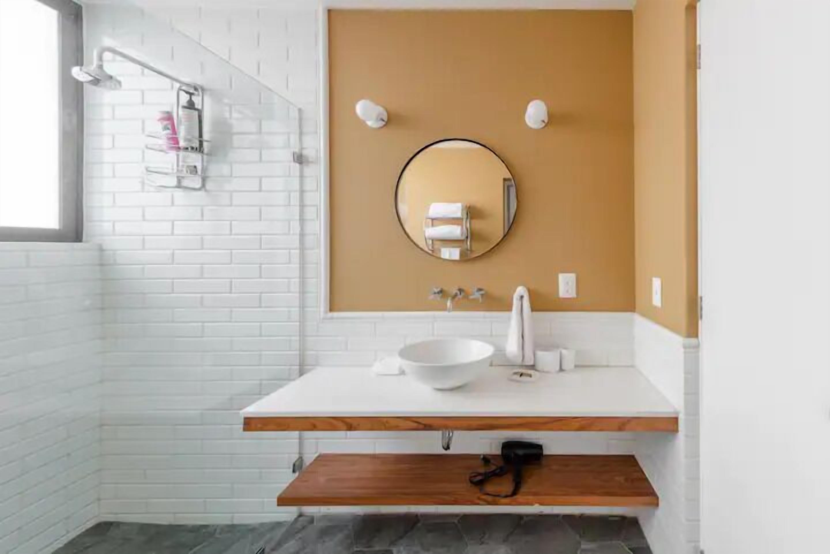 A modern bathroom with a white sink, wooden shelf, and a round mirror on the wall.