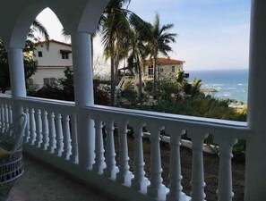 A balcony with a white balustrade overlooking a coastal view with palm trees and a building with a red roof. - Peaceful Ocean View Studio  (Puerto Plata)