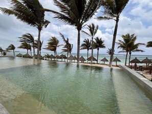 A pool area with palm trees and thatched umbrellas, overlooking the ocean.
