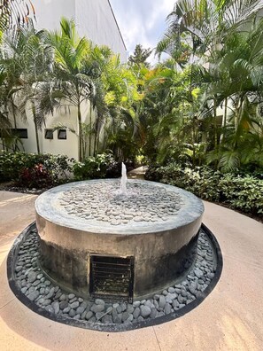 Una fuente de piedra circular con agua brotando del centro, rodeada por un anillo de piedras más pequeñas y un camino empedrado.