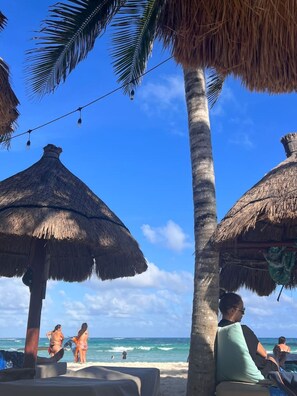 Escena de playa con sombrillas de paja, palmeras y un cielo azul despejado.
