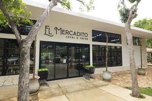 Fachada de un comercio llamado 'El Mercado Local & Fresh', con grandes ventanales de vidrio y plantas en macetas afuera.