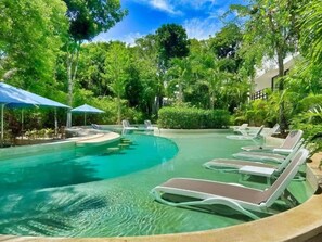 Una piscina con sillones de descanso y sombrillas, rodeada de abundante vegetación.