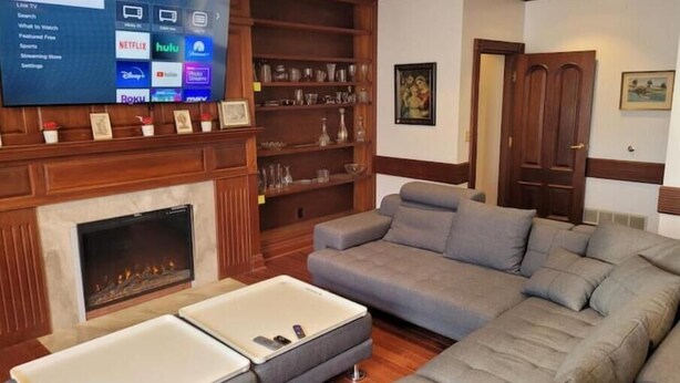 Una sala de estar con un televisor de pantalla plana grande mostrando la interfaz de un servicio de streaming, una chimenea, un sofá seccional y una estantería de madera.