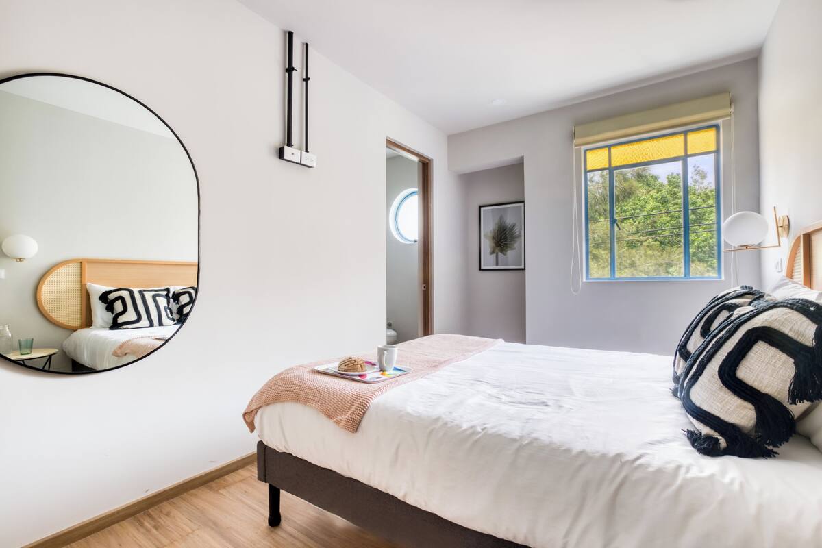 Un dormitorio con una cama grande, un espejo redondo, una mesita de noche, una ventana con vistas a la vegetación y una puerta que da a otra habitación.