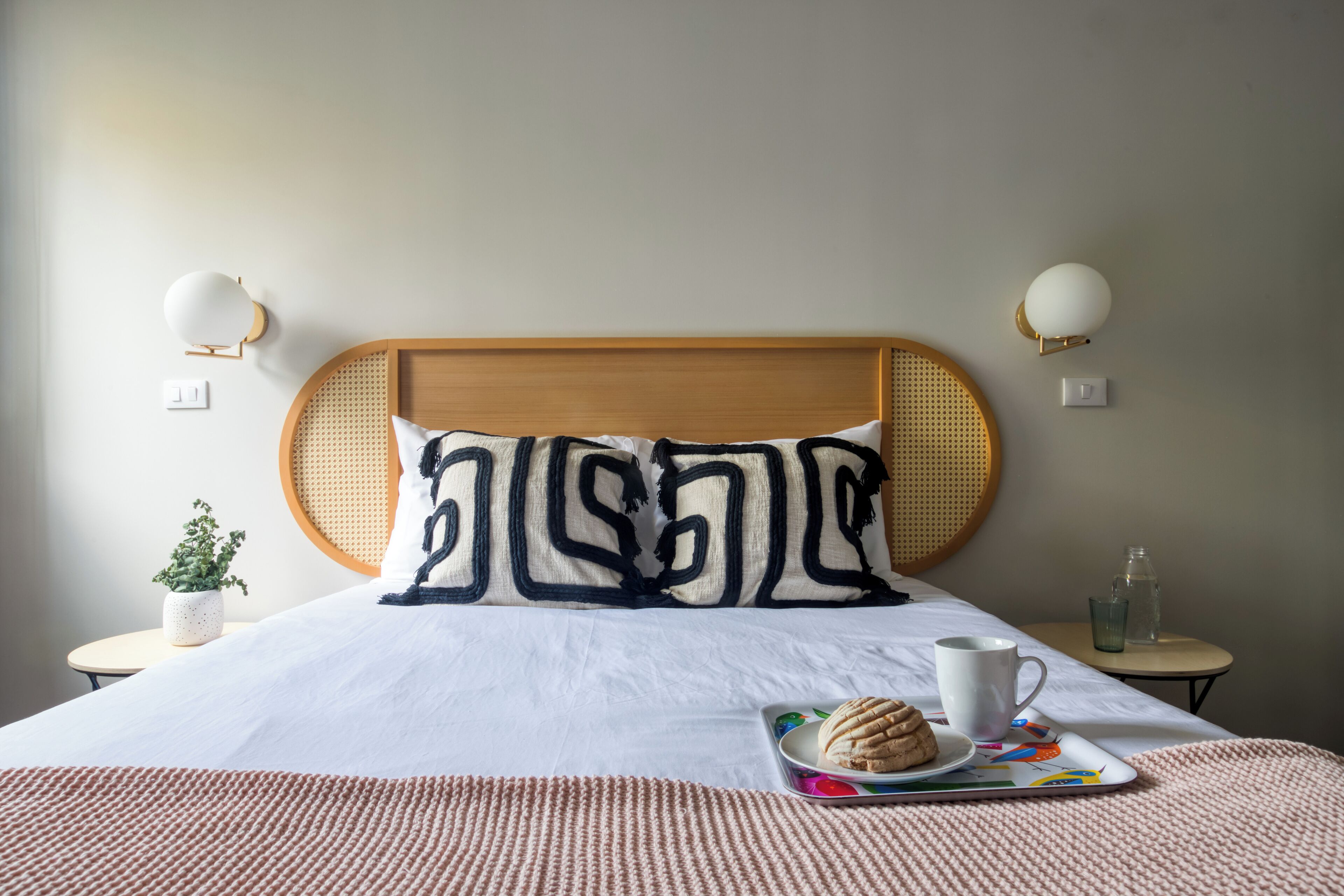 Un lit bien fait, recouvert d’une couette blanche, avec un coussin décoratif, un plateau sur lequel se trouvent une pâtisserie et une tasse, et une petite table avec un verre d’eau.
