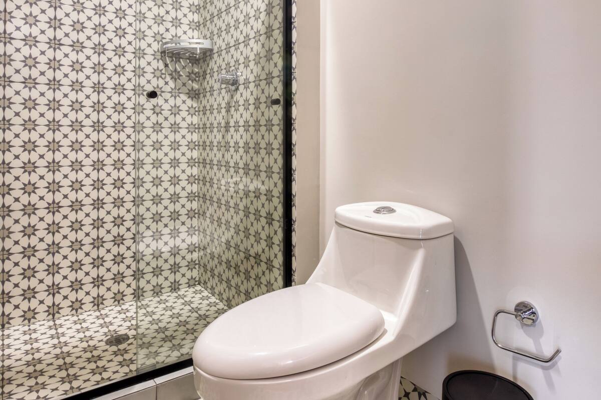 Un baño con inodoro, cabina de ducha con azulejos decorativos y un cubo de basura.