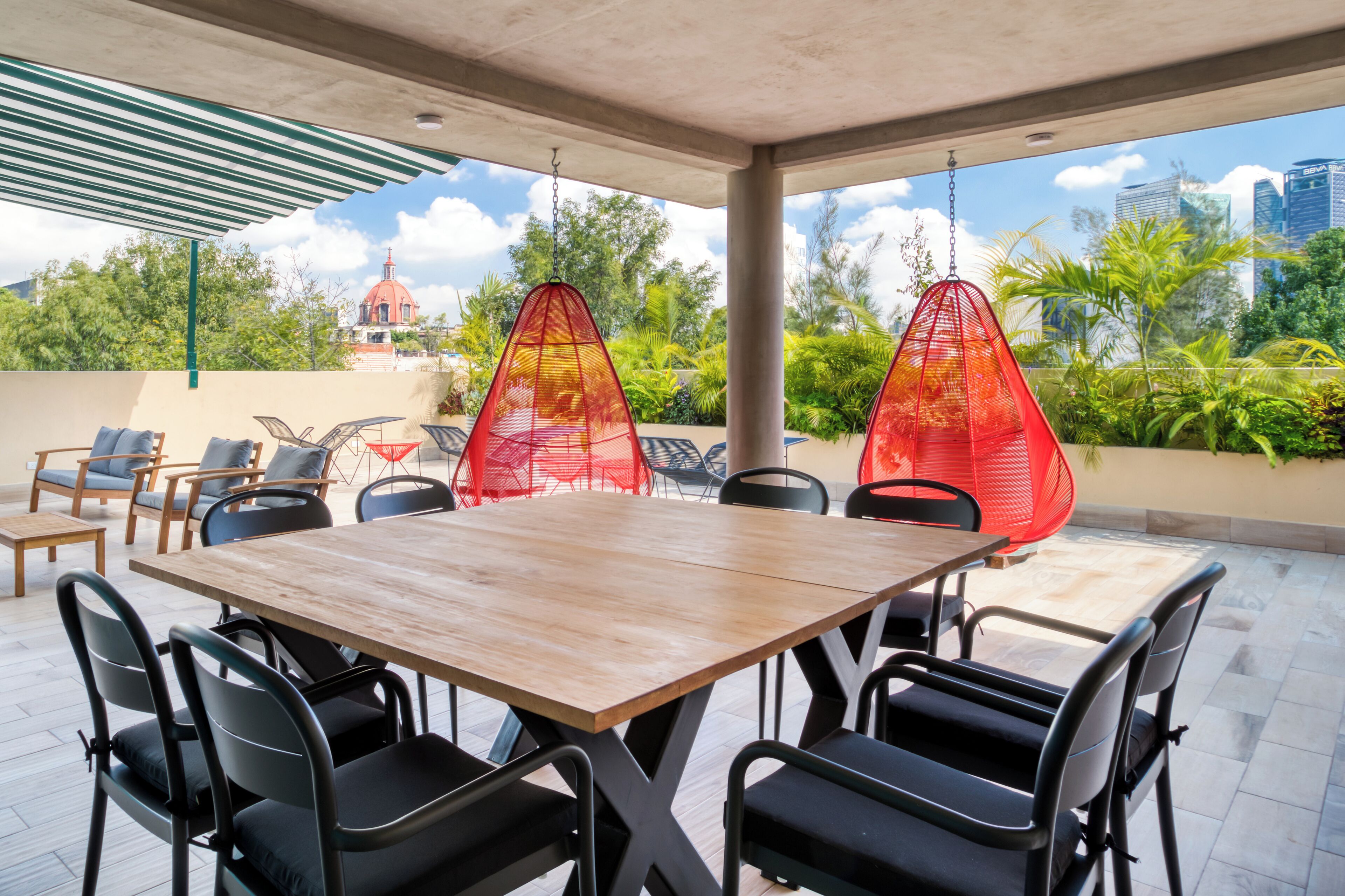 An outdoor patio with a wooden table and chairs, red hanging chairs, and a view of a cityscape and greenery.