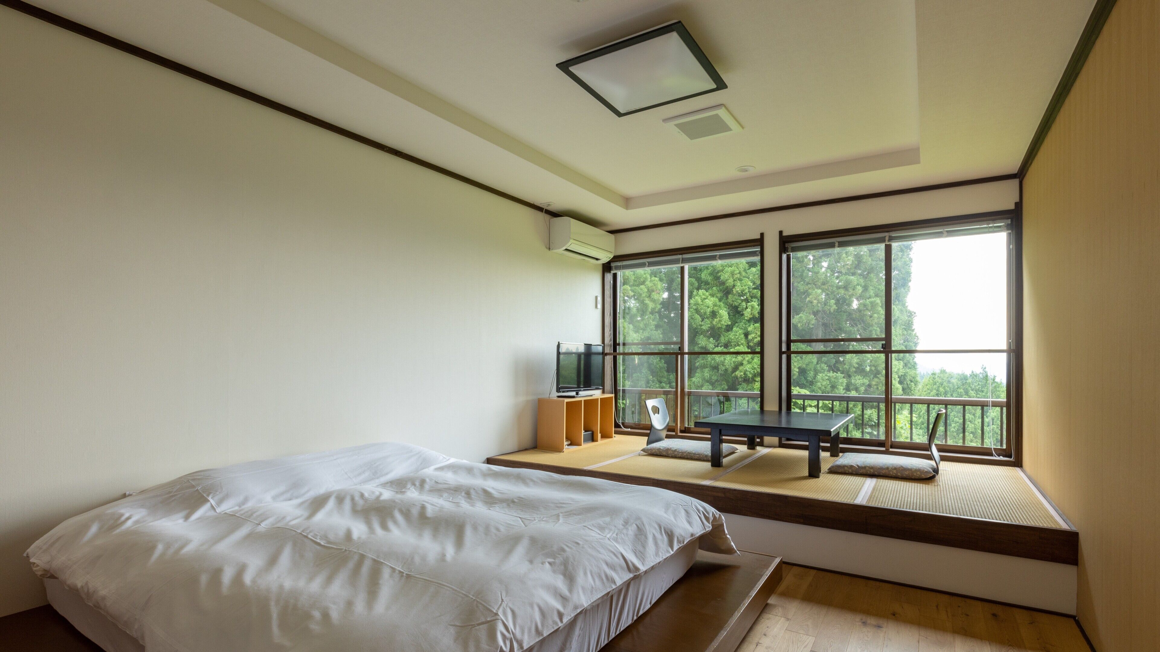 A bedroom with a bed, a TV, a small table, and a window with a view of trees.