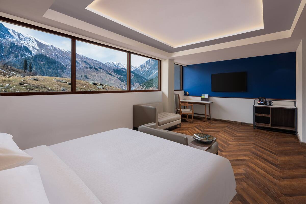 Quarto de hotel com uma cama grande, uma mesa, uma cadeira e uma televisão. O quarto tem vista para as montanhas através de janelas amplas.
