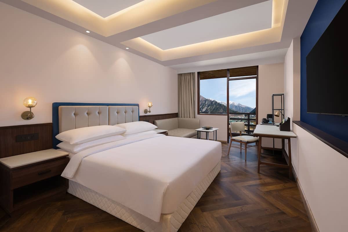 Quarto de hotel com uma cama grande, uma escrivaninha, uma cadeira e vista para montanhas através de uma janela ampla.