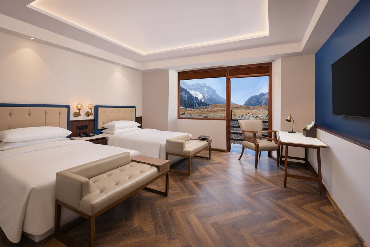Quarto de hotel com duas camas, uma escrivaninha, uma cadeira e uma grande janela com vista para as montanhas.