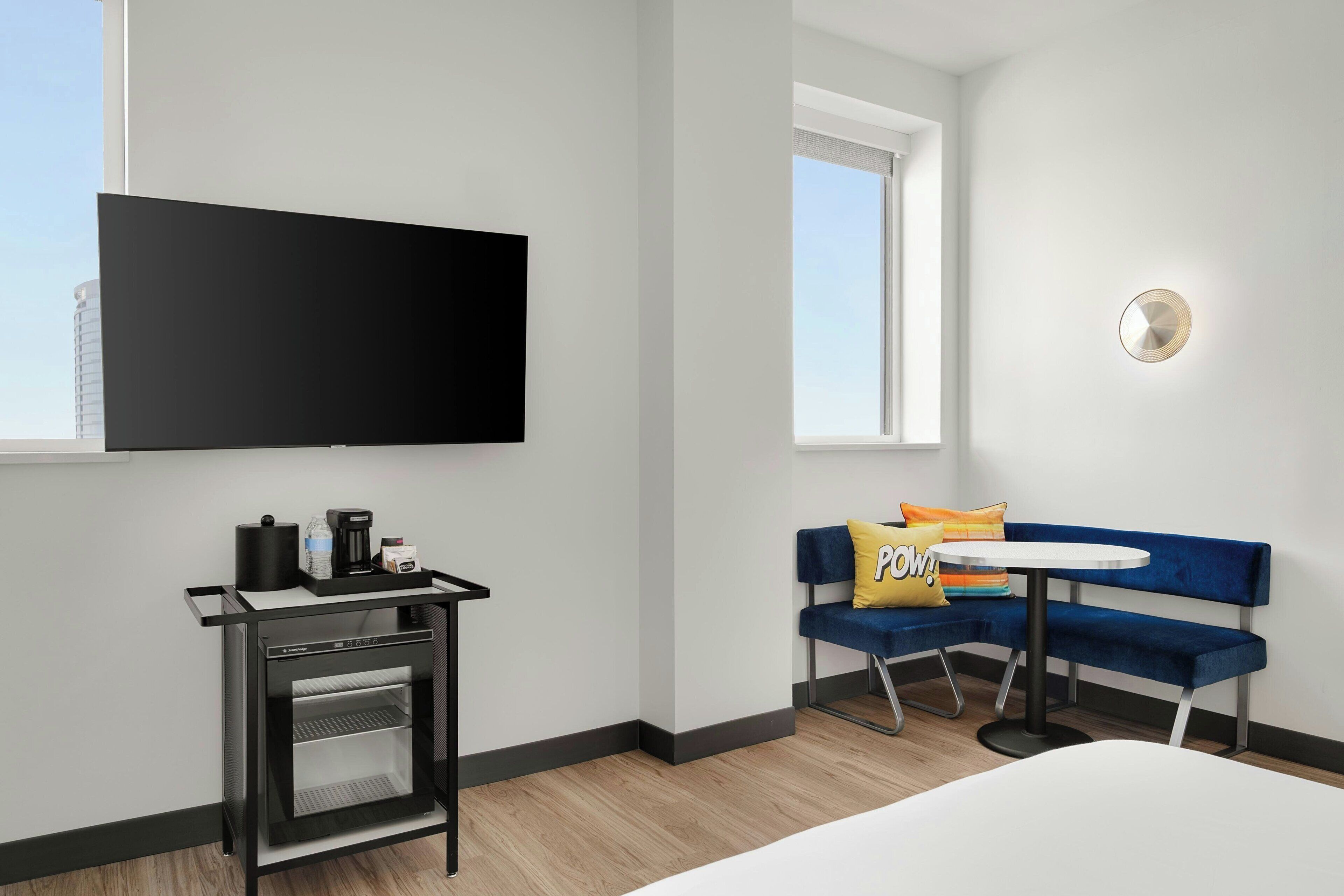 A modern hotel room with a flat-screen TV, a small kitchenette, a blue sofa with a 'POW!' pillow, and a round table.