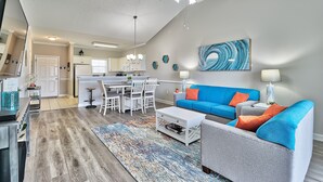 Ein modernes Wohnzimmer mit einem blauen Sofa, einem grauen Sessel und einem Couchtisch. Im Hintergrund befindet sich eine Küche mit weißen Schränken und ein Essbereich mit weißen Stühlen.