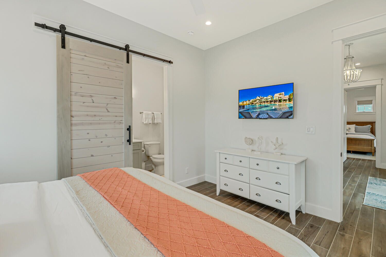 A bedroom with a bed, a white dresser, a painting of a beach scene, and a bathroom visible through an open door.
