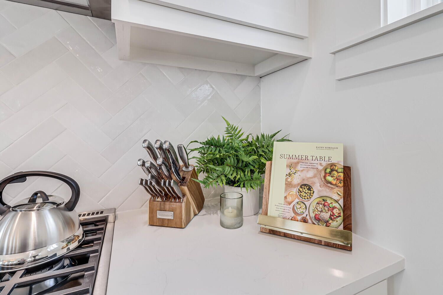 A kitchen with a kettle, knife block, potted plant, and a cookbook titled 'Summer Table'.