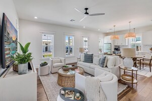 A spacious living room with a sofa, coffee table, and a dining area with chairs. There is a kitchen visible in the background.