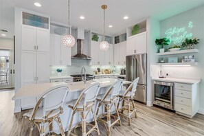 A modern kitchen with white cabinetry, a central island with bar stools, stainless steel appliances, and a neon sign on the wall. - Ships and Giggles 276 HI (Port Aransas)