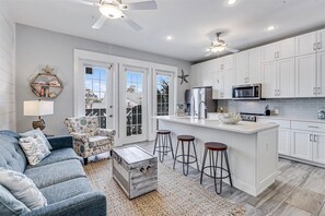 A modern kitchen with white cabinets, a central island with stools, a ceiling fan, and a view through large windows.