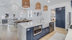 A modern kitchen with a large island, double sink, and built-in oven. There are wicker pendant lights, a dining area with a table and chairs, and a view into the adjacent living room. - Sunnyside Up 209 LAK (Port Aransas)