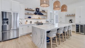 A modern kitchen with white cabinetry, a stainless steel refrigerator, a marble island, wicker pendant lights, and a coffee machine. - Sunnyside Up 209 LAK (Port Aransas)