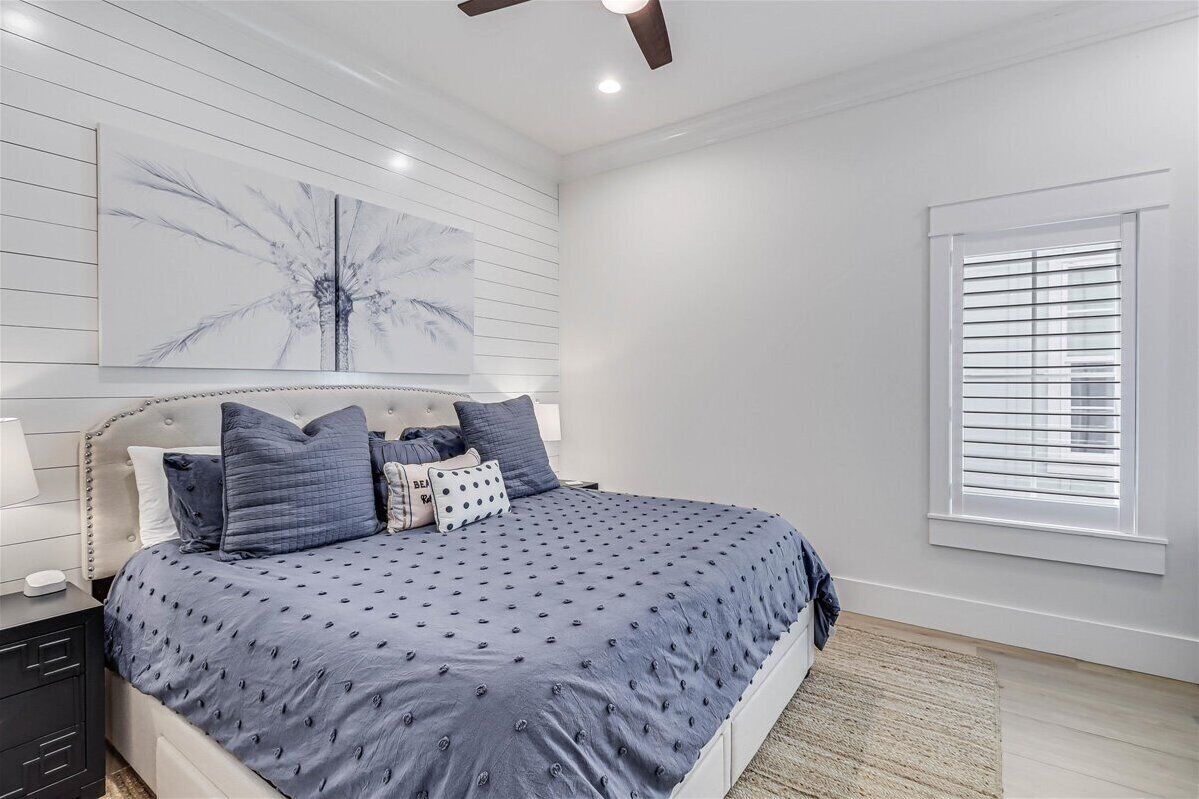 A bedroom with a large bed, a ceiling fan, a window with blinds, and a wall-mounted artwork.
