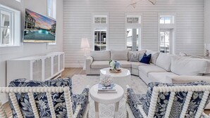 A spacious living room with a white sofa, matching armchairs, a coffee table, and a painting of a sunset over a pool. - Sunnyside Up 209 LAK (Port Aransas)