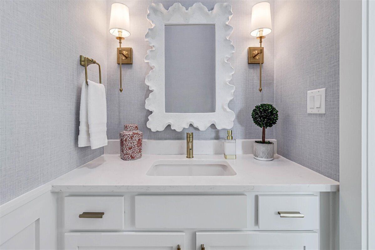 Un baño con tocador blanco, un espejo grande con marco ondulado, dos luces empotradas en la pared, un grifo dorado, un lavabo blanco, una pequeña planta en maceta y un jarrón decorativo.