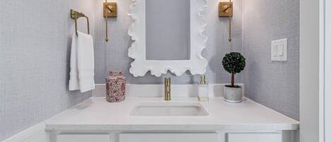 Un baño con tocador blanco, un espejo grande con marco ondulado, dos luces empotradas en la pared, un grifo dorado, un lavabo blanco, una pequeña planta en maceta y un jarrón decorativo.